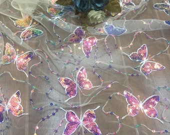 May include: A sheer, off-white tulle fabric featuring an all-over pattern of iridescent, multi-coloured sequined butterflies. The butterflies are connected by thin, delicate lines of small, colourful sequins. Three artificial fabric roses are placed on top.
