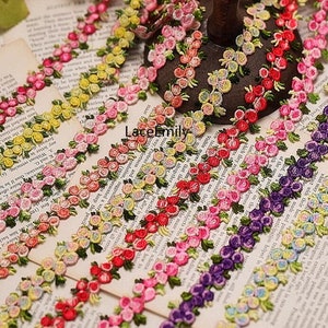 Può includere: Una varietà di pizzo floreale ricamato colorato con un motivo ripetitivo di piccoli fiori e foglie. Il pizzo è disposto in file su uno sfondo bianco.