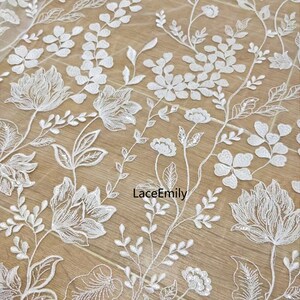 Pu&ograve; includere: Tessuto di pizzo floreale bianco con un delicato motivo di fiori e foglie. Il tessuto &egrave; trasparente e ha un leggero luccichio. Il testo "LaceEmily" &egrave; visibile nella parte inferiore centrale dell'immagine.