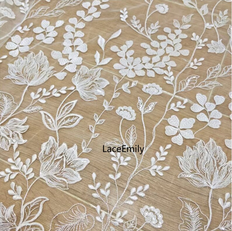 Pu&ograve; includere: Tessuto di pizzo floreale bianco con un delicato motivo di fiori e foglie. Il tessuto &egrave; trasparente e ha un leggero luccichio. Il testo "LaceEmily" &egrave; visibile nella parte inferiore centrale dell'immagine.