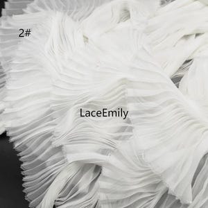 May include: A white pleated fabric trim featuring a dense, layered ruffle design. The material is semi-sheer with fine, vertical folds throughout. The text 2# and LaceEmily are printed in black on the surface of the white fabric.