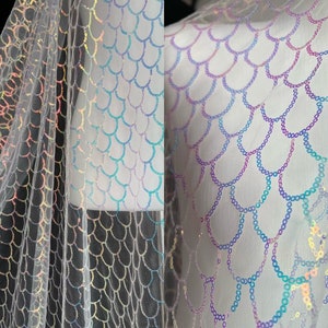 May include: White iridescent mermaid scale fabric with a mesh backing. The scales are made of sequins and are arranged in a pattern that resembles fish scales.