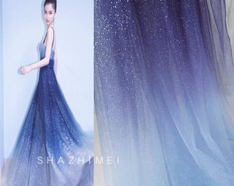 May include: A woman wearing a long, flowing dress with a blue gradient, transitioning from dark navy to light blue. The dress has a shimmering, glittery finish. The brand name "SHAZHIMEI" is displayed on the dress.