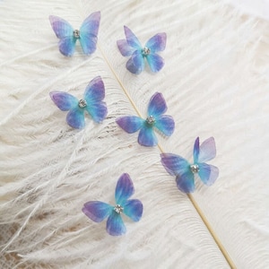 May include: Six blue and purple butterfly embellishments with clear rhinestone centers. The butterflies are made of sheer fabric and are arranged on a white feather background.
