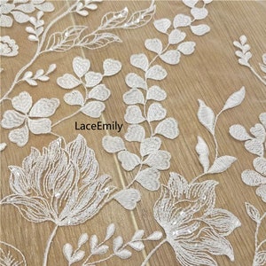 Pu&ograve; includere: Tessuto di pizzo floreale bianco con un delicato motivo di foglie e fiori. Il tessuto &egrave; impreziosito da piccole paillettes, che aggiungono un tocco di brillantezza.