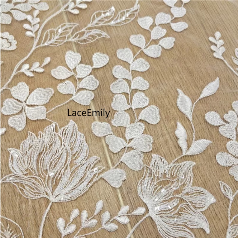 Pu&ograve; includere: Tessuto di pizzo floreale bianco con un delicato motivo di foglie e fiori. Il tessuto &egrave; impreziosito da piccole paillettes, che aggiungono un tocco di brillantezza.