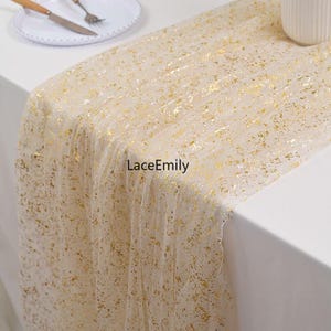 May include: A sheer, cream-colored table runner with gold foil accents. The runner is draped over a white table, with a plate, knife, and fork visible in the background. The text "LaceEmily" is printed on the runner.