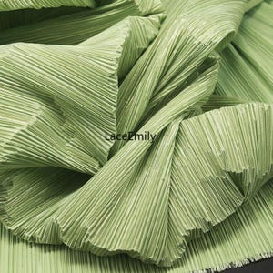 May include: Close-up of a light green pleated fabric. The fabric is folded and draped, showcasing the texture and pattern of the pleats. The fabric has a subtle sheen, and the folds create shadows and highlights.