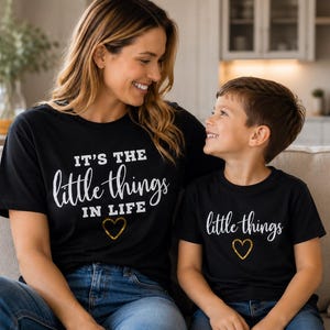 Puede incluir: Camisetas negras con el texto "It's the little things in life" y "little things" en escritura blanca, con un corazón dorado. Las camisetas las llevan una mujer y un niño. Están sentados en un sofá beige.