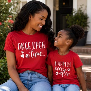 Könnte beinhalten: Zwei Personen tragen passende rote T-Shirts mit weißem Text. Ein Shirt sagt "ONCE UPON a time..." und das andere sagt "HAPPILY ever after" mit Pfotenabdrücken. Beide lächeln.