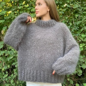 Könnte beinhalten: Ein grauer, übergroßer Pullover mit hohem Ausschnitt und langen Ärmeln. Der Pullover hat ein weiches, strukturiertes Aussehen und besteht aus einem dicken Strickmaterial. Das Model trägt eine weiße Hose. Der Hintergrund ist ein grüner, belaubter Busch.
