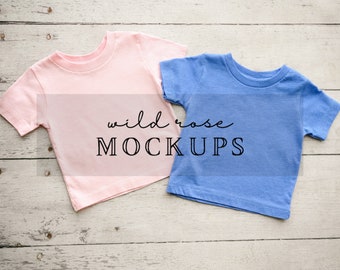 Op de afbeelding: Twee blanco T-shirts, een roze en een blauwe, plat gelegd op een witgeschilderd houten oppervlak. De tekst "wild rose MOCKUPS" is onder de shirts gedrukt.