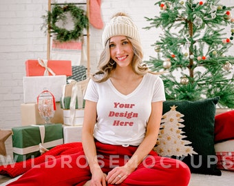Puede incluir: Una mujer con una camiseta blanca de cuello en V con el texto "Your Design Here" en letras rojas. Lleva una boina de punto blanca y pantalones rojos. Está sentada frente a un árbol de Navidad y regalos.