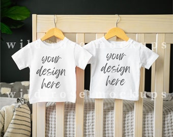Puede incluir: Dos bodies blancos para bebé colgados de perchas de madera en una cuna blanca. Los bodies tienen el texto "your design here" impreso en ellos.