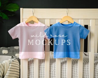 Op de afbeelding: Twee T-shirts met korte mouwen en een ronde hals, één lichtroze en één blauw, hangend aan houten kledinghangers. De tekst "wild rose MOCKUPS" staat in het wit over het midden van beide shirts, die voor een licht houten frame hangen.