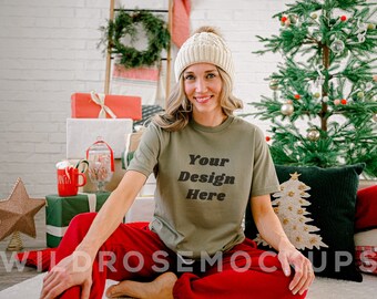 Puede incluir: Una mujer con una camiseta verde con el texto "Your Design Here" en negro. Está sentada en una alfombra blanca con pantalones rojos y una boina de punto blanca. Hay decoraciones navideñas en el fondo.