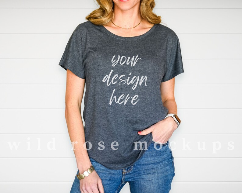 May include: A dark charcoal grey short-sleeved t-shirt with a scoop neckline. The front of the shirt features the white text "your design here" in a cursive script font. The shirt is paired with blue denim jeans and a white smartwatch on the wrist.