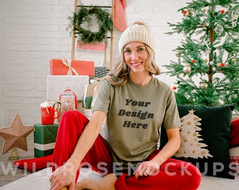 Puede incluir: Una mujer con una camiseta verde con el texto "Your Design Here" en letras negras. Lleva una boina de punto blanca y pantalones rojos. Está sentada frente a un árbol de Navidad y regalos.