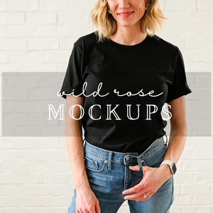 以下が含まれることがあります： 白いレンガの壁の前に立っている女性。黒いTシャツを着ており、Tシャツには「wild rose MOCKUPS」の文字が書かれている。青いジーンズを履いている。