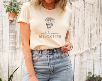 以下が含まれることがあります： 白と黒の骸骨とバラのグラフィックと「wild rose MOCKUPS」のテキストが書かれた薄いベージュのTシャツを着た女性。彼女は青いデニムのショートパンツを履いています。