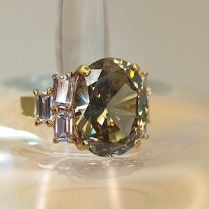 Op de afbeelding: Een gouden ring met een grote, ovaal geslepen, champagnekleurige edelsteen als middelpunt. Kleinere, rechthoekige, lichtgekleurde stenen flankeren de hoofdsteen. De ring is gezet op een gouden band.