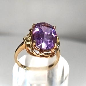 May include: A gold ring with a large, oval-shaped purple gemstone. The gemstone is set in an ornate gold setting. The ring's band is a simple, polished gold. The ring is a piece of jewellery.