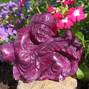 Pode incluir: Uma escultura esculpida de uma figura sentada em um tom magenta profundo. A figura usa um chapéu e vestes, com uma expressão sorridente. A escultura está colocada sobre uma base de pedra, com vibrantes flores rosa e roxas ao fundo.