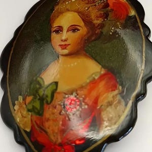 May include: An oval-shaped black brooch featuring a hand-painted portrait of a person with an elaborate hairstyle adorned with a red feather. The figure wears a peach-coloured garment with a green bow, a pearl necklace, and a prominent red brooch.