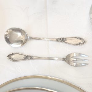 May include: Silverware set featuring a soup spoon and a fork with three tines. Both pieces have ornate, decorative detailing on the handles. The silverware is placed on a white tablecloth, with a portion of a plate visible.