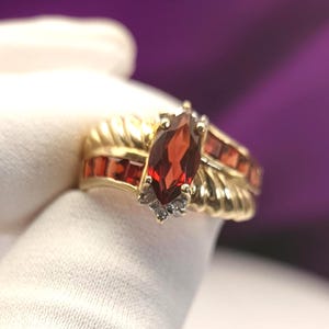 Può includere: Un anello in oro con una pietra granato rosso scuro taglio marquise al centro, affiancata da granati rossi taglio quadrato incastonati a canale lungo la fascia. Piccole pietre chiare sono incastonate sotto il granato centrale e la fascia è scanalata.