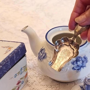 May include: A white ceramic teapot featuring blue floral patterns and a blue rim, accompanied by a silver-toned metal tea scoop with an ornate engraved handle and a scalloped bowl, positioned near a rectangular tin with a blue and white floral design.
