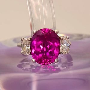 May include: A silver ring with a large, oval magenta gemstone and two smaller, clear stones. The ring's design is set against a blurred background. The focus is on the vibrant colours and the intricate details of the jewellery.