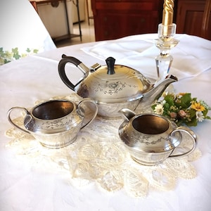 Könnte beinhalten: Ein silbernes Teeservice mit Teekanne, Sahnekännchen und Zuckerdose, präsentiert auf einer weißen Spitzendecke. Die Teekanne hat einen schwarzen Griff und einen Deckelknopf. Blumendesigns sind auf dem Silber geätzt.