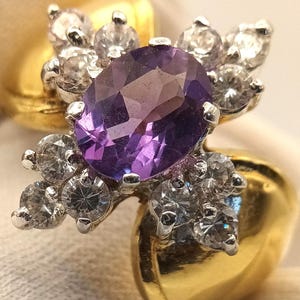 Op de afbeelding: Een gouden ring met een grote, ovaal geslepen paarse edelsteen, omringd door kleinere, heldere edelstenen. Het ontwerp van de ring omvat clusters van heldere stenen aan weerszijden van de centrale steen, wat een sprankelende, elegante uitstraling creëert. De ring is gezet op een gouden band.