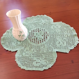 Peut inclure: Un vase en céramique blanc avec un design cannelé et des accents de trèfle est posé sur un napperon en crochet vert clair. Le napperon a une forme de trèfle à quatre feuilles avec un motif ajouré détaillé. Les articles sont présentés sur une surface en bois.
