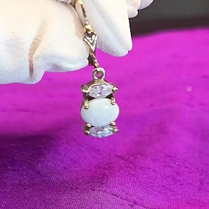 Könnte beinhalten: Ein goldfarbener Ohrring mit einem Bügelverschluss. Der Ohrring hat einen weißen, oval geformten Stein, der von zwei klaren, marquiseförmigen Steinen flankiert wird. Der Ohrring wird vor einem lila Hintergrund präsentiert.