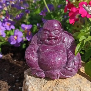 May include: A carved, smiling Buddha statue in a deep magenta hue. The seated figure is on a light brown stone, surrounded by purple and pink flowers. The Buddha's form is detailed, with visible folds and a serene expression.