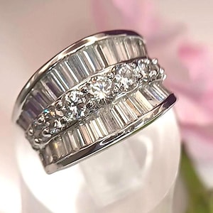 May include: A silver-toned ring featuring a central row of five round-cut clear stones flanked by two rows of baguette-cut clear stones. The band is wide and metallic, showcasing a multi-row design with a polished finish and clear, faceted stones.