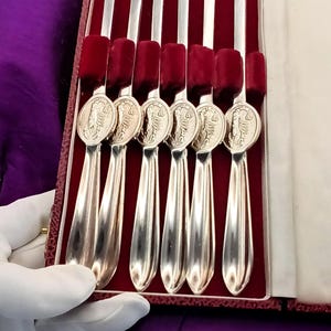 May include: A set of six silver-toned metal cocktail forks with two-pronged tips and oval medallions featuring embossed animal figures on the handles, presented in a rectangular case with a deep red velvet interior lining and white trim.