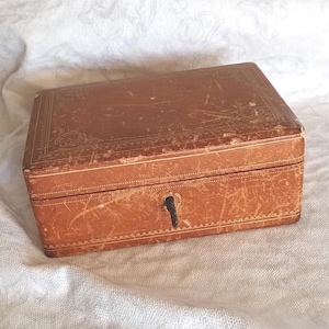 Könnte beinhalten: Eine rechteckige Box aus braunem Leder mit goldfarbenen, geprägten Zierrändern und Mustern auf dem Deckel und den Seiten. Die Box verfügt über ein zentrales Schlüsselloch an der Vorderseite mit einem kleinen, dunkelmetallenen Schlüssel.