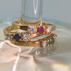 Può includere: Tre anelli d'oro con pietre preziose. Un anello presenta pietre chiare e rosse, un altro ha pietre chiare, blu e chiare, e il terzo anello ha pietre chiare e verdi. Gli anelli sono impilati.