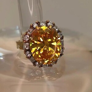 May include: A gold-coloured ring with a large, oval-cut yellow gemstone as its centrepiece. The gemstone is encircled by smaller, clear, rectangular-cut stones and gold accents. The ring's band is a simple gold colour.