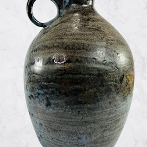 Puede incluir: Una jarra de cerámica hecha a mano con una superficie oscura y texturizada. La jarra presenta un cuerpo redondeado, un cuello estrecho y un asa en forma de lazo. La paleta de colores incluye tonos de azul, gris y marrón.