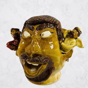 Op de afbeelding: Een geglazuurde keramische pot in de vorm van een menselijk hoofd met een bruine baard en haar, grote witte ogen en een open mond. Een klein rood figuurtje is aan de linkerkant bevestigd en een klein geel gevleugeld figuurtje aan de rechterkant.