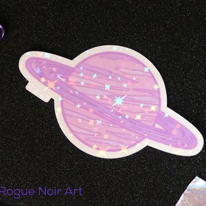 K&ouml;nnte beinhalten: Ein holografischer Aufkleber mit einem rosa Planeten mit einem Ring darum. Der Planet ist mit kleinen wei&szlig;en Sternen bedeckt. Der Aufkleber befindet sich auf einem schwarzen Hintergrund.