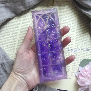 May include: A rectangular, transparent resin bookmark with a purple, starry design. The bookmark features a window-like pattern with arched tops and a glittery, celestial aesthetic. The text "Rogue Noir Art" is visible.