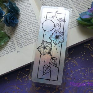 May include: A holographic bookmark with a black border featuring a moon and a vine with flowers. The background is a rainbow gradient.