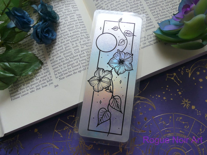 May include: A holographic bookmark with a black border featuring a moon and a vine with flowers. The background is a rainbow gradient.