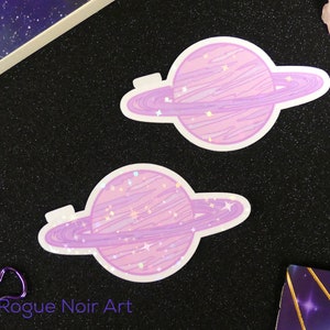 K&ouml;nnte beinhalten: Zwei violette und rosafarbene Glitzer-Planeten-Aufkleber mit Ringen. Die Planeten haben eine glitzernde, holografische Oberfl&auml;che.