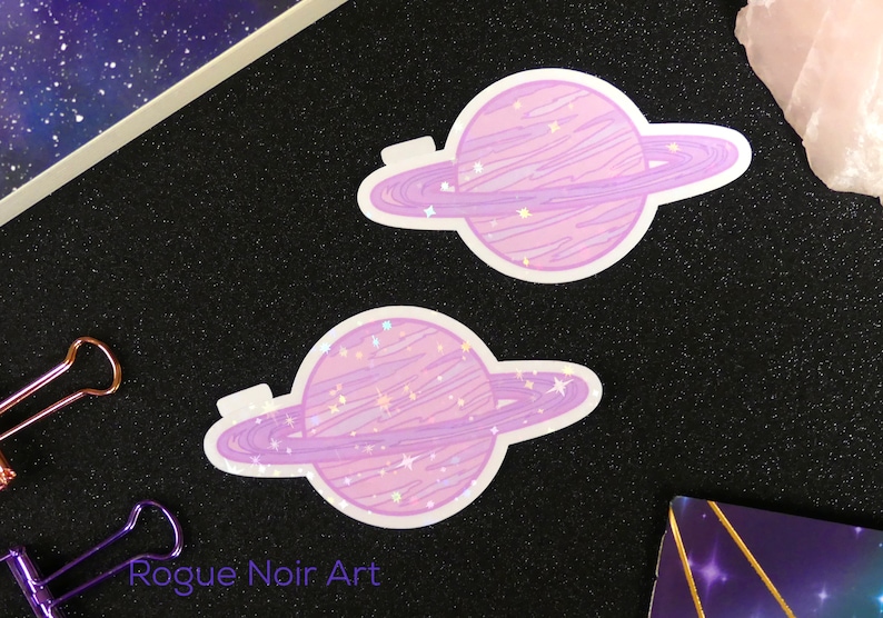 K&ouml;nnte beinhalten: Zwei violette und rosafarbene Glitzer-Planeten-Aufkleber mit Ringen. Die Planeten haben eine glitzernde, holografische Oberfl&auml;che.